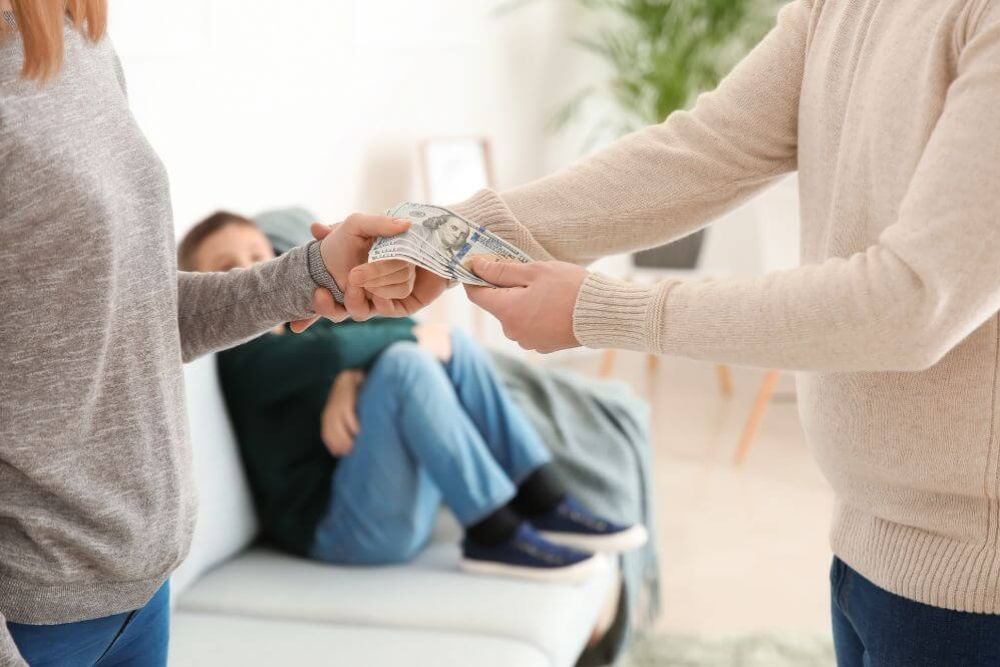A person in a gray sweater hands a stack of cash to another person in a beige sweater while a child in a dark green shirt sits on a couch, observing.