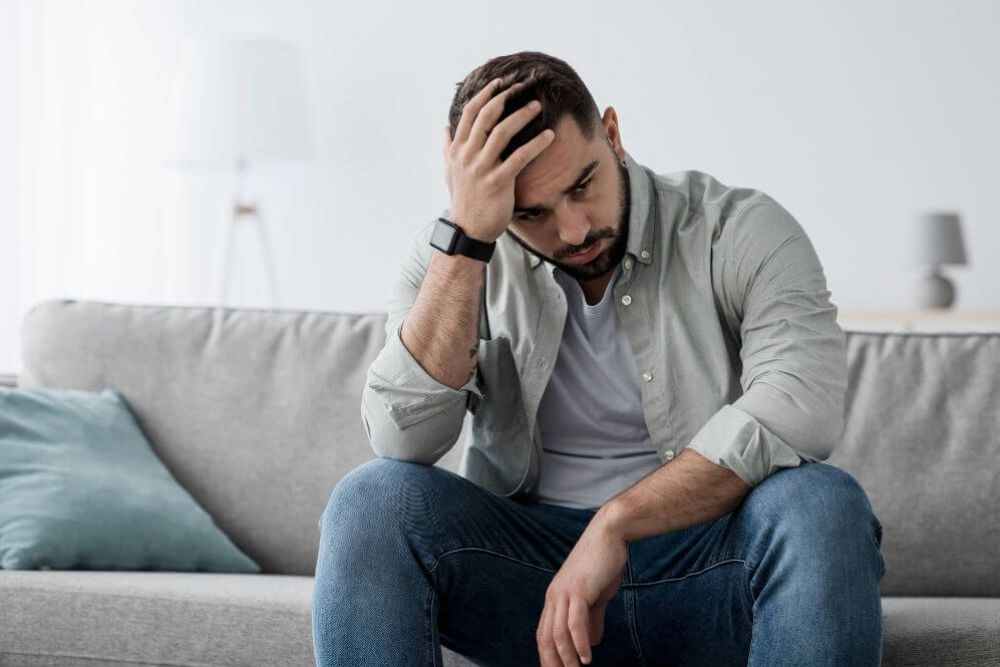 A man sits on a sofa, resting his head on his hand, looking pensive or troubled. He is in a modern, minimalistic living room with soft lighting.