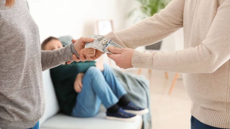 A person in a gray sweater hands a stack of cash to another person in a beige sweater while a child in a dark green shirt sits on a couch, observing.