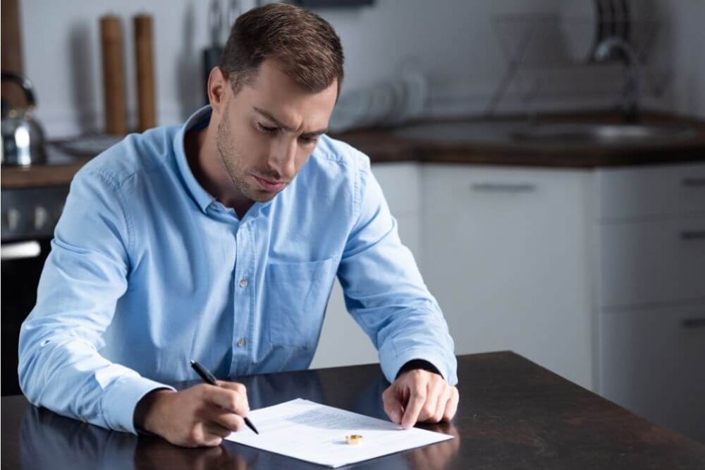 A man in a blue shirt is writing on a piece of paper seated at a wooden table in a modern kitchen, showcasing a focused expression while contemplating his task.