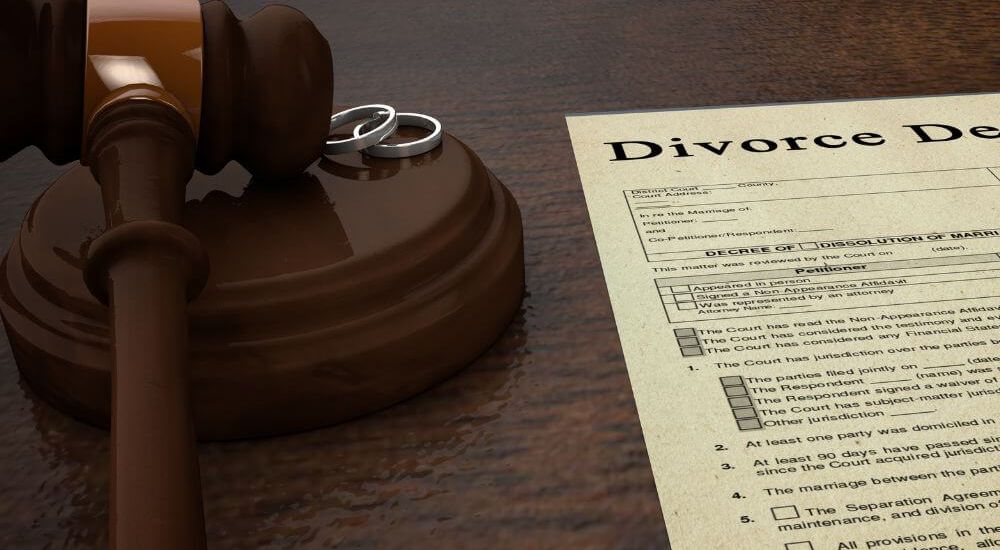 A gavel rests on a wooden surface next to two wedding rings and a divorce decree document. The context conveys a legal setting, signifying the end of a marriage.