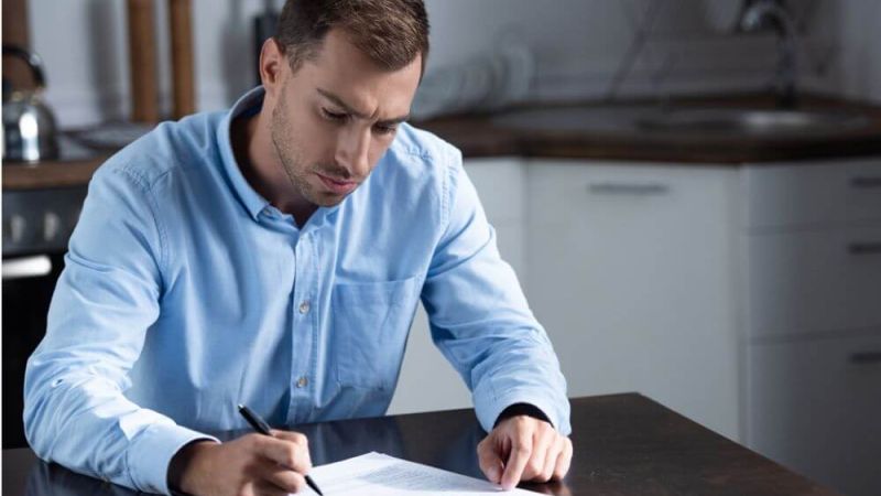 A man in a blue shirt is writing on a piece of paper seated at a wooden table in a modern kitchen, showcasing a focused expression while contemplating his task.