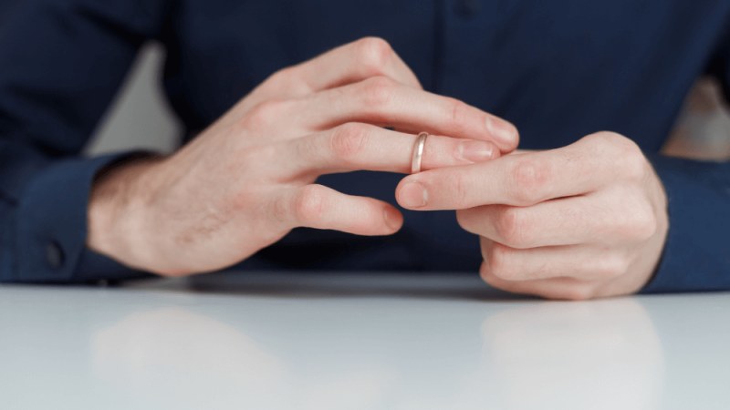 A hand with a simple ring is shown in the process of being placed on another finger, set against a light-colored tabletop, with the individual wearing a dark blue shirt.