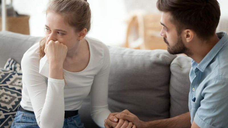 A woman sits on a sofa, appearing thoughtful and concerned, with her hand over her mouth, while a man holds her other hand in a supportive gesture.