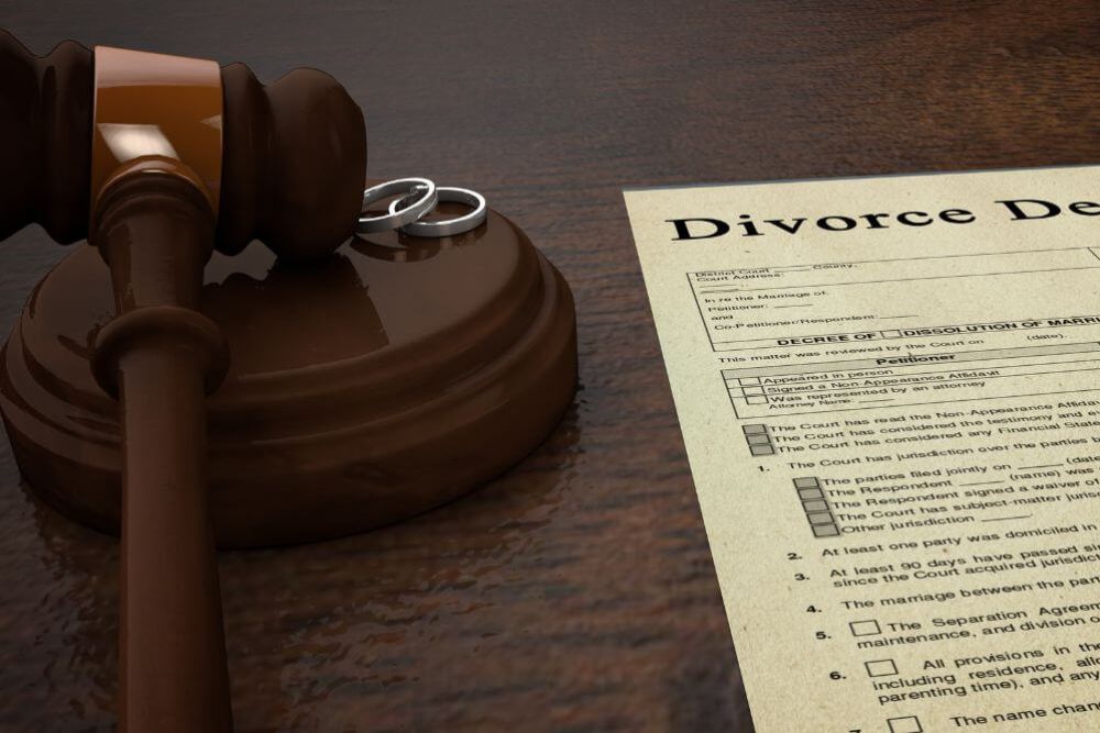 A gavel rests on a wooden surface next to two wedding rings and a divorce decree document. The context conveys a legal setting, signifying the end of a marriage.